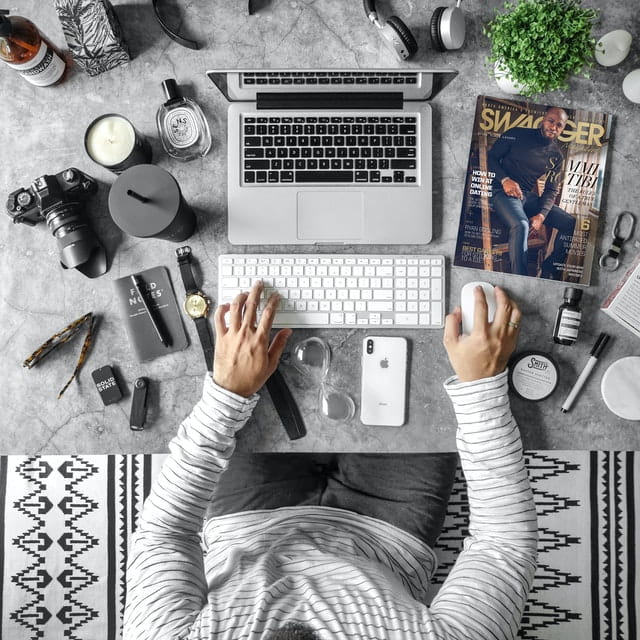 A person working on a laptop surrounded by various objects on a marble desk with a black and white rug underneath