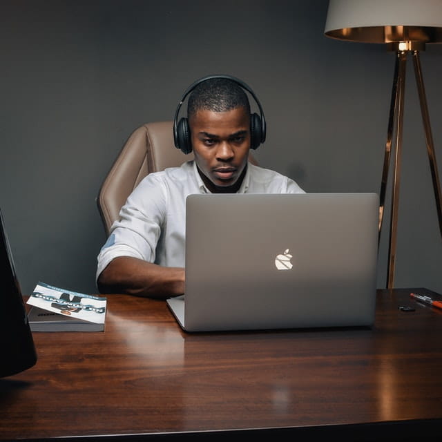 A person wearing headphones and working on a silver Macbook in a home office with a wooden desk, lamp, magazine, and pen